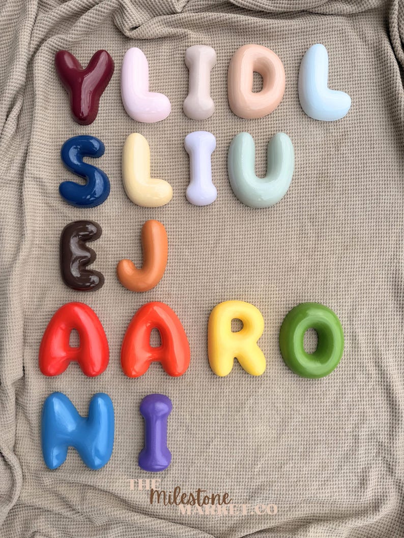 May include: A collection of twenty-one individual, three-dimensional, glossy, balloon-style letters in various colours including red, blue, yellow, green, orange, purple, brown, and pastel shades. The text "THE Milestone MARKET CO" is printed at the bottom.