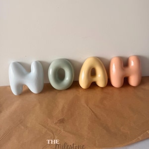 May include: Four individual ceramic letters spelling the name NOAH in a rounded, bubble-like font. The letters are coloured in light blue, sage green, soft yellow, and peach. The text THE MILESTONE MARKET.CO is printed in white at the bottom.