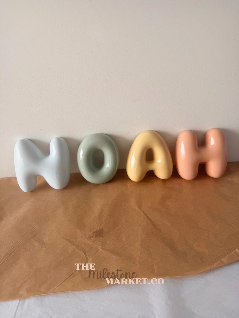 May include: Four individual ceramic letters spelling the name NOAH in a rounded, bubble-like font. The letters are coloured in light blue, sage green, soft yellow, and peach. The text THE MILESTONE MARKET.CO is printed in white at the bottom.