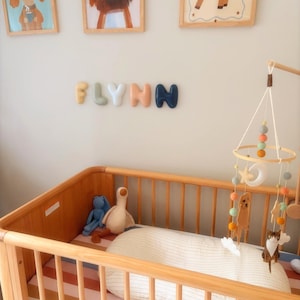 May include: A wooden cot with a striped mattress, a white pillow, and a hanging mobile featuring felt animals, stars, and a moon. On the wall above, the name FLYNN is spelled out in individual, rounded, multi-coloured 3D letters.