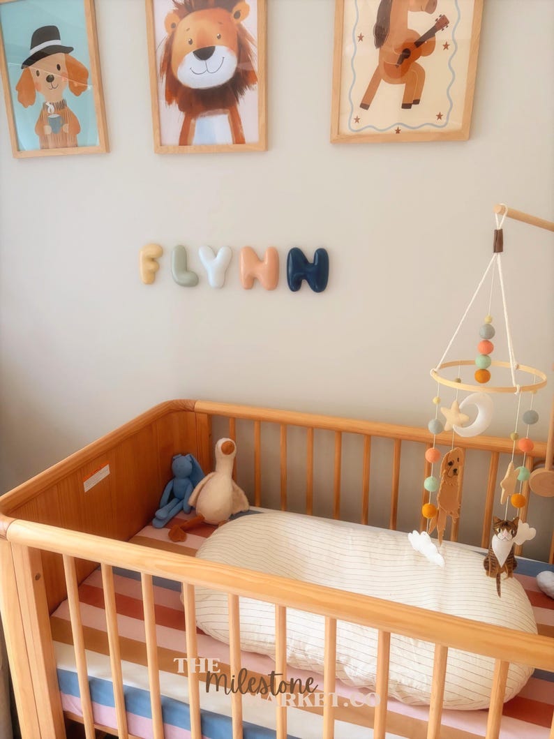 May include: A wooden cot with a striped mattress, a white pillow, and a hanging mobile featuring felt animals, stars, and a moon. On the wall above, the name FLYNN is spelled out in individual, rounded, multi-coloured 3D letters.