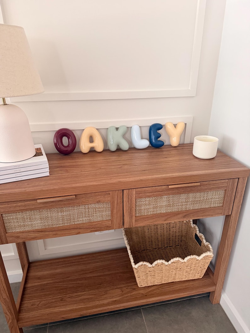 May include: A set of individual, rounded, three-dimensional letters spelling the name OAKLEY in various colours including dark red, light yellow, sage green, light blue, dark blue, and peach, displayed on a wooden console table.