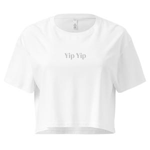 Könnte beinhalten: Ein weißes, gecropptes T-Shirt mit kurzen Ärmeln. Der Text "Yip Yip" ist in Hellgrau auf die Brust gedruckt. Das Shirt hat einen Rundhalsausschnitt und eine lockere Passform, geeignet für Freizeitkleidung.