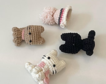 Op de afbeelding: Vier handgemaakte gehaakte items: een bruine hond, een witte kat, een zwarte kat en een roze en witte hoed met een pompon. De honden- en katfiguren hebben roze sjaals en zwarte knoopogen. De hoed heeft roze, grijze en witte strepen.