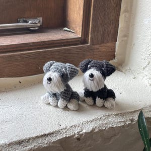 Könnte beinhalten: Zwei handgefertigte Schnauzer-Hundefiguren aus Häkelarbeit. Einer ist grau und weiß, der andere schwarz und weiß. Beide haben weiße Bärte und Augenbrauen sowie schwarze Knopfaugen. Sie sitzen auf einer weißen Oberfläche.