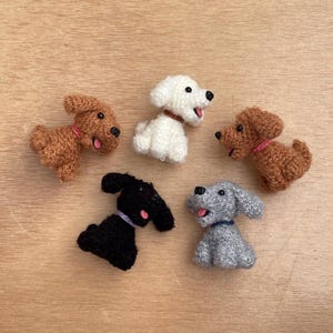 Op de afbeelding: Vijf handgemaakte gehaakte hondenfiguurtjes in bruin, wit, zwart en grijs. Elke hond heeft een halsband en een roze of blauwe tong. De honden zijn gerangschikt op een lichtbruin houten oppervlak.