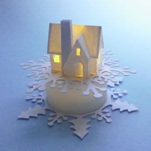 May include: A white paper house with a lit window, sitting on a white paper snowflake.