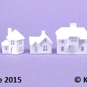 May include: A row of nine white paper model houses of varying sizes, arranged from smallest to largest. The houses are all simple in design, with no windows or doors visible. The houses are all white and have a slightly textured surface. The text "Micro sized Tiny Village 2015" is at the bottom left of the image and the text "© Karin Corbin 2015" is at the bottom right of the image.