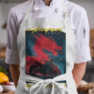 Puede incluir: Delantal blanco con un gr&aacute;fico de Cyberpunk 2077, que muestra un drag&oacute;n rojo y una persona en una motocicleta. El delantal tiene un dise&ntilde;o de lazo y el texto "WAKE UP SAMURAI". Llevado por una persona con una chaqueta de chef blanca.