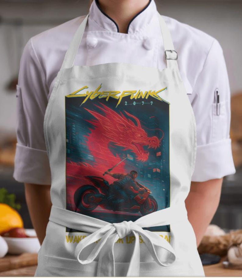 Puede incluir: Delantal blanco con un gr&aacute;fico de Cyberpunk 2077, que muestra un drag&oacute;n rojo y una persona en una motocicleta. El delantal tiene un dise&ntilde;o de lazo y el texto "WAKE UP SAMURAI". Llevado por una persona con una chaqueta de chef blanca.