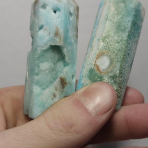 May include: Two blue-green larimar crystal towers. The crystals have a rough, textured surface with brown and white inclusions. One tower has a carved-out section. The crystals are held in a hand.