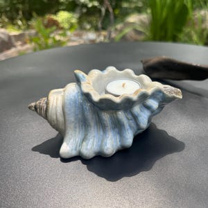 May include: A ceramic seashell-shaped candle holder with a glossy blue and white glaze finish. The shell features a central circular opening containing a small white tea light candle. A piece of dark brown driftwood is positioned nearby.