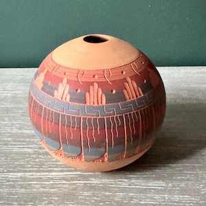 May include: A spherical terracotta vase featuring a small circular opening at the top. The surface is decorated with etched geometric patterns in shades of dark gray and reddish-brown, including repeating Greek key motifs and vertical lines.
