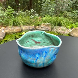 May include: A hand-thrown ceramic bowl with an irregular, wavy rim. The exterior features a blend of deep blue and turquoise glazes with vertical drip patterns. The interior is finished in a vibrant green glaze with visible brown clay undertones.