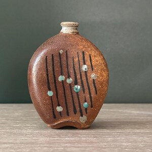 May include: A small, flattened, round ceramic vase with a textured, mottled brown glaze. The front surface features vertical black lines interspersed with small, raised, circular dots in shades of light blue, white, and grey. It has a narrow neck.