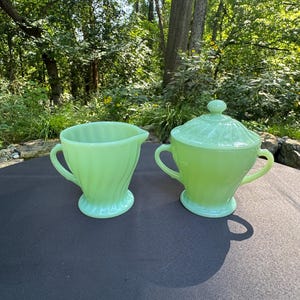 May include: A light green jadeite creamer and a matching lidded sugar bowl. Both pieces feature a swirling ribbed pattern, a pedestal base, and curved handles. The sugar bowl has a removable lid with a small knob handle on the top center.