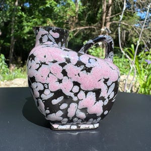 May include: A ceramic pitcher with a black base and handle, adorned with a pink and white floral pattern. The pitcher has a curved body, a spout, and a sturdy handle. The design is a mix of abstract and natural elements.