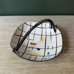 May include: A shallow, square-shaped bowl with rounded corners, featuring a cream-colored base with a black grid pattern. The bowl includes small, scattered rectangular accents in yellow, blue, and brown, and a black, textured, arched handle.