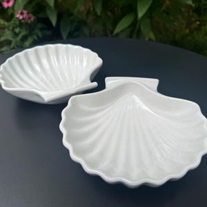 May include: Two white ceramic dishes shaped like scallop shells. Each dish features a glossy finish with raised, radiating ridges that mimic the natural texture of a seashell, creating a scalloped edge along the perimeter of the bowl.