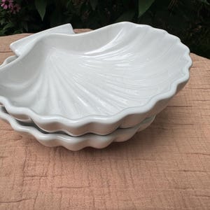 May include: A stack of two white ceramic seashell-shaped dishes. The dishes have a scalloped edge and a textured, fan-like pattern on the inside. The dishes are sitting on a light brown surface.
