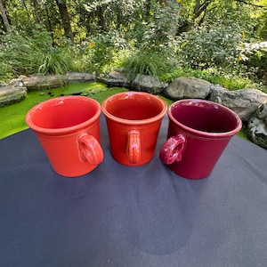 May include: Three ceramic mugs arranged in a row, featuring a smooth, glossy finish. The mug on the left is bright orange, the center mug is a vibrant red, and the mug on the right is a deep, dark magenta. Each mug has a sturdy, curved handle.