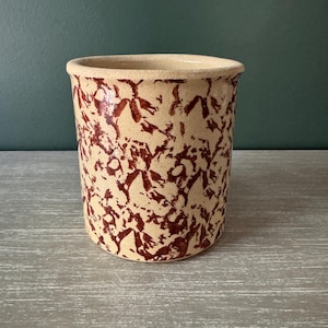May include: A cylindrical ceramic crock featuring a cream-colored base with a dark reddish-brown sponge-painted or mottled pattern throughout the exterior surface. The vessel has a thick, rounded rim and a wide, open top with a smooth interior.
