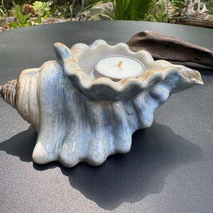 May include: A ceramic candle holder shaped like a conch shell with a glossy finish in shades of light blue, white, and tan. The shell features a scalloped opening that holds a single round white tea light candle with a small visible wick.
