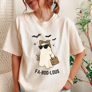 May include: Ivory-colored t-shirt with a cartoon ghost wearing sunglasses, a leopard print bow, and carrying a coffee cup and a leopard print bag. The shirt has the text "FA-BOO-LOUS" and three bat graphics. The shirt is a Halloween-themed design.