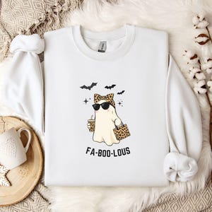 May include: White sweatshirt with a Halloween-themed graphic. The design features a ghost wearing sunglasses and a leopard print bow, holding a coffee cup and a leopard print bag. The text "FA-BOO-LOUS" is below the ghost.