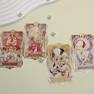 Könnte beinhalten: Vier dekorative Metallkarten im Jugendstil mit goldenen Rahmen. Jede Karte zeigt eine andere Illustration mit leuchtenden Farben, darunter Rot, Rosa, Lila und Gold. Die Karten sind auf einer hellgrünen Oberfläche mit verstreuten Perlen angeordnet.
