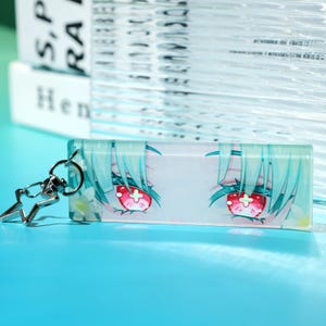 May include: A rectangular acrylic keychain featuring a graphic illustration of anime-style eyes with mint green hair, bright red irises with white cross symbols, and small white flowers, attached to a silver-colored star-shaped metal clasp.