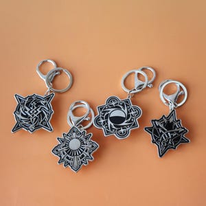 Puede incluir: Cinco llaveros plateados con intrincados diseños geométricos en negro y plata. Cada llavero tiene una forma diferente, incluyendo explosiones de estrellas y patrones abstractos. Fondo naranja.