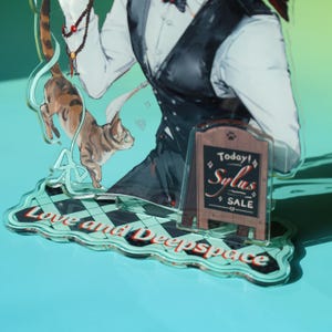 May include: An acrylic stand featuring a character in a white shirt and black vest, with a cat illustration. The stand has a sign that says "Today! Sylus SALE" and the words "Love and Deepspace". The stand is on a blue surface.