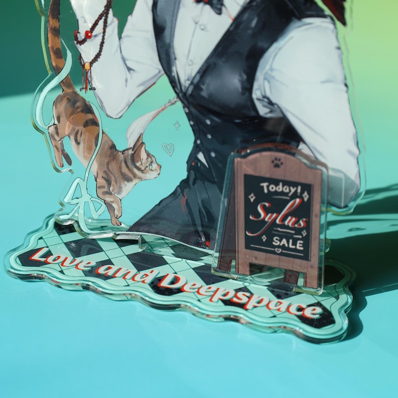 May include: An acrylic stand featuring a character in a white shirt and black vest, with a cat illustration. The stand has a sign that says "Today! Sylus SALE" and the words "Love and Deepspace". The stand is on a blue surface.
