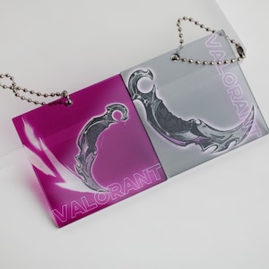 Puede incluir: Dos llaveros cuadrados con un diseño de cuchillo karambit. Uno es magenta con la palabra "VALORANT", el otro es gris con la palabra "VALORANT". Ambos tienen una cadena de bolas plateada.