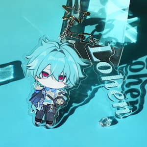 Puede incluir: Un llavero de acrílico transparente con un personaje de anime estilo chibi de cabello azul claro y ojos rojos, con un atuendo azul y blanco. Un dije rectangular de acrílico unido al broche en forma de estrella muestra el nombre Lohen.