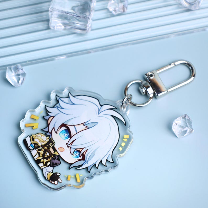 May include: A clear acrylic keychain featuring a cartoon character with white hair and blue eyes. The keychain has a silver clasp and a decorative design. The background is light blue with scattered clear crystals.
