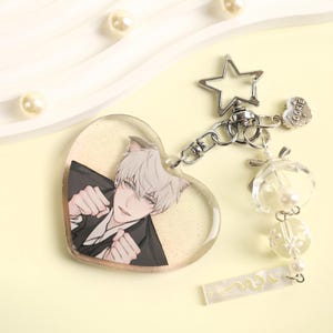 Op de afbeelding: Een hartvormige sleutelhanger met een anime-achtig personage met wit haar en kattenoren, gekleed in een zwart pak. De sleutelhanger heeft een zilveren ster en een bedel met het woord "COOKIE". Extra bedels zijn heldere kralen en parels.
