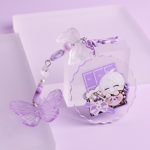 Könnte beinhalten: Ein lilafarbener Schlüsselanhänger mit einem Schmetterlingsanhänger und einem kreisförmigen Anhänger mit einer Cartoonfigur. Der Schmetterling ist durchscheinend mit detaillierten Flügelmustern. Der kreisförmige Anhänger hat einen gewellten Rand und eine Cartoon-Illustration.