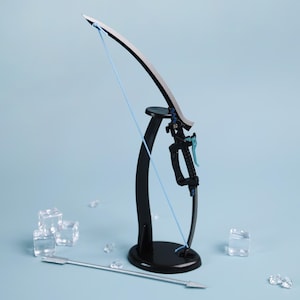 May include: A black and silver bow and arrow set with a blue string. The bow is mounted on a black stand. An arrow lies on the surface. The background is a light blue color.