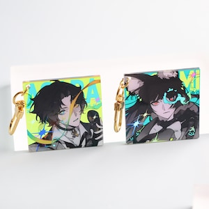 Puede incluir: Dos llaveros cuadrados con arte de personajes estilo anime. Uno presenta un personaje masculino con cabello negro y acentos amarillos, el otro un personaje femenino con cabello negro y ojos rojos. Ambos tienen cierres dorados.