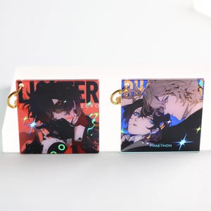 Op de afbeelding: Twee vierkante sleutelhangers met goudkleurige ringen. Eén sleutelhanger heeft een rode achtergrond met een donkerharig anime-personage en de tekst "LUSTER ZERO". De andere sleutelhanger heeft een blauwe achtergrond met twee anime-personages en de tekst "PHAETHON".