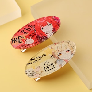 May include: Two oval-shaped hair clips with anime-style character designs. One clip is red with a character illustration and text, the other is yellow with a different character and text. Both clips have silver metal clasps.