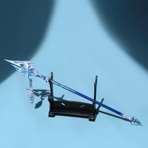May include: A blue and white fantasy spear with intricate detailing, displayed on a black stand against a blue backdrop. The spear features a decorative head and a hanging charm, showcasing a blend of fantasy and craftsmanship.