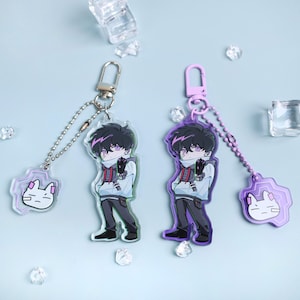 Peut inclure: Deux porte-clés de style anime représentant un personnage aux cheveux noirs, un sweat à capuche blanc et un pantalon gris. Les porte-clés ont une chaîne argentée et violette et un charm en forme de chat. Le fond est bleu clair.