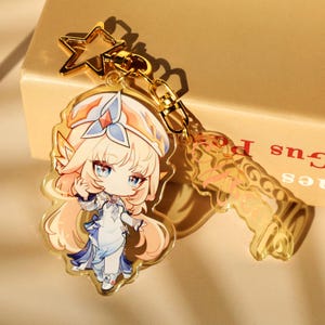 Puede incluir: Un llavero de acrílico con un personaje estilo chibi de cabello rubio largo, que viste un atuendo blanco y azul con un sombrero a juego. El dije está sujeto a un broche dorado en forma de estrella e incluye una etiqueta de acrílico.