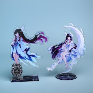 Puede incluir: Dos soportes acrílicos con personajes de estilo anime con pelo largo negro y atuendos fluidos azules y blancos. Un personaje está de pie, el otro está sentado en una luna creciente. El primer soporte tiene la palabra "Columbina" en la base.