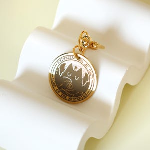 May include: A gold-colored keychain with a round pendant. The pendant features a stylized cat design with the text "LORD OF THE MYSTERIES" around the edge. The keychain is on a white, wavy surface.