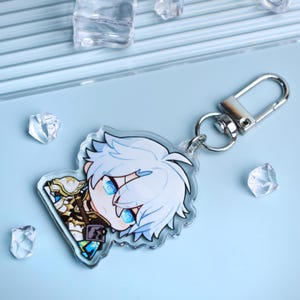 May include: A clear acrylic keychain featuring a cartoon character with white hair and blue eyes. The keychain has a silver clasp and a small metal ring. The character is wearing a detailed outfit with gold and brown accents. The background is light blue.