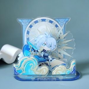 May include: A clear acrylic standee featuring a chibi anime character with light blue hair and attire. The character is set against a backdrop of a celestial design with the word "PHAINON" at the base. The standee has a detailed, layered design.